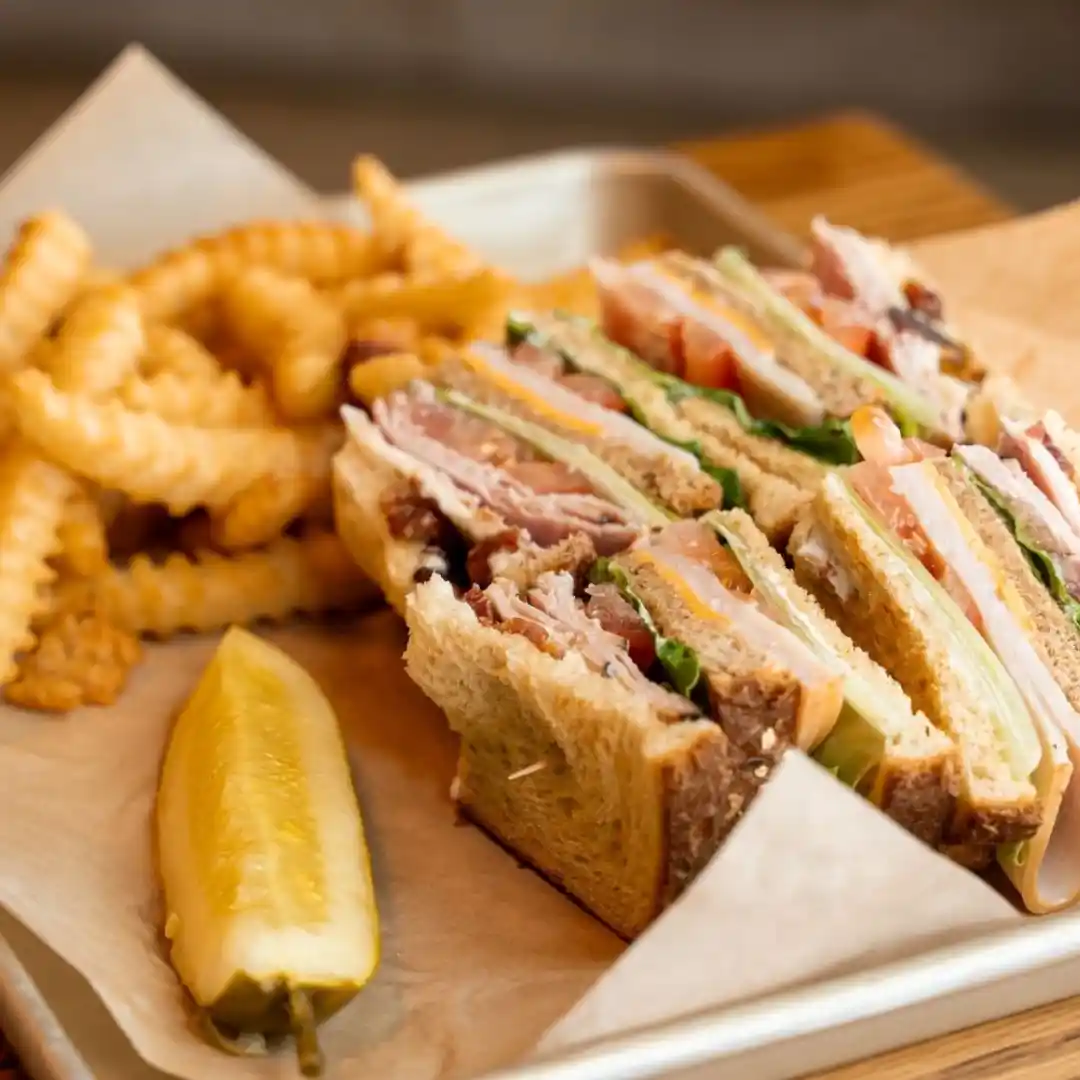 A tray with a club sandwich cut into four triangles, stacked with turkey, bacon, lettuce, and tomato, served with crinkle-cut fries and a pickle spear on parchment paper.