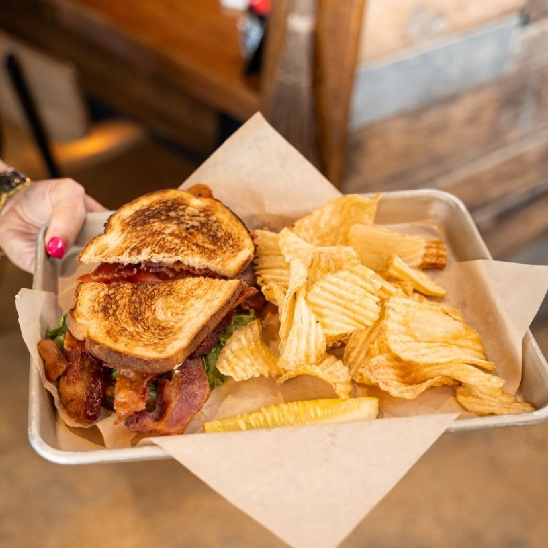 A hand holds a tray with a BLT sandwich on toasted bread, a pile of ridged potato chips, and a pickle spear, all served on brown parchment paper.