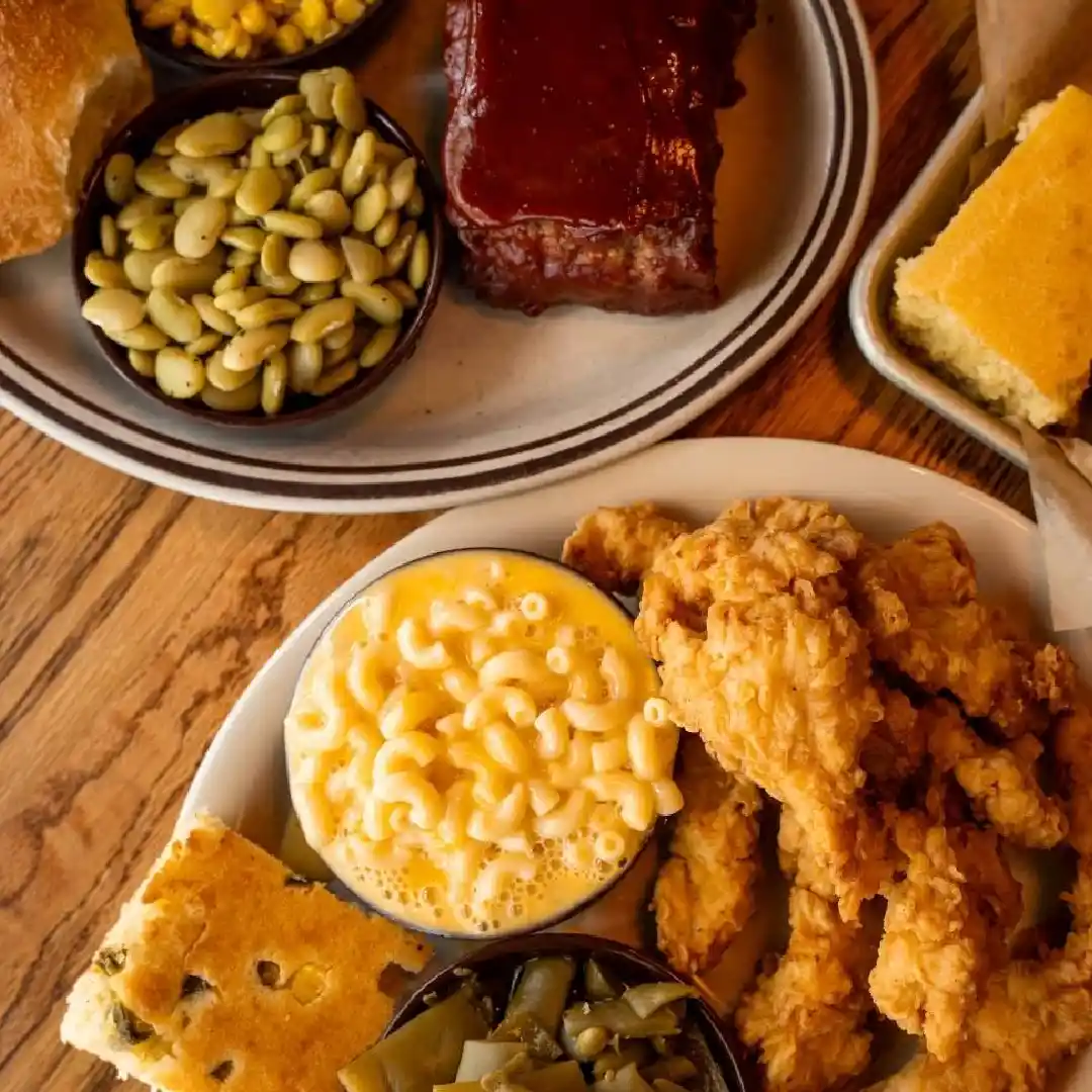 Two plates of Southern comfort food, including fried chicken, macaroni and cheese, green beans, cornbread, butter beans, corn, and a slice of barbecue meatloaf on a wooden table.