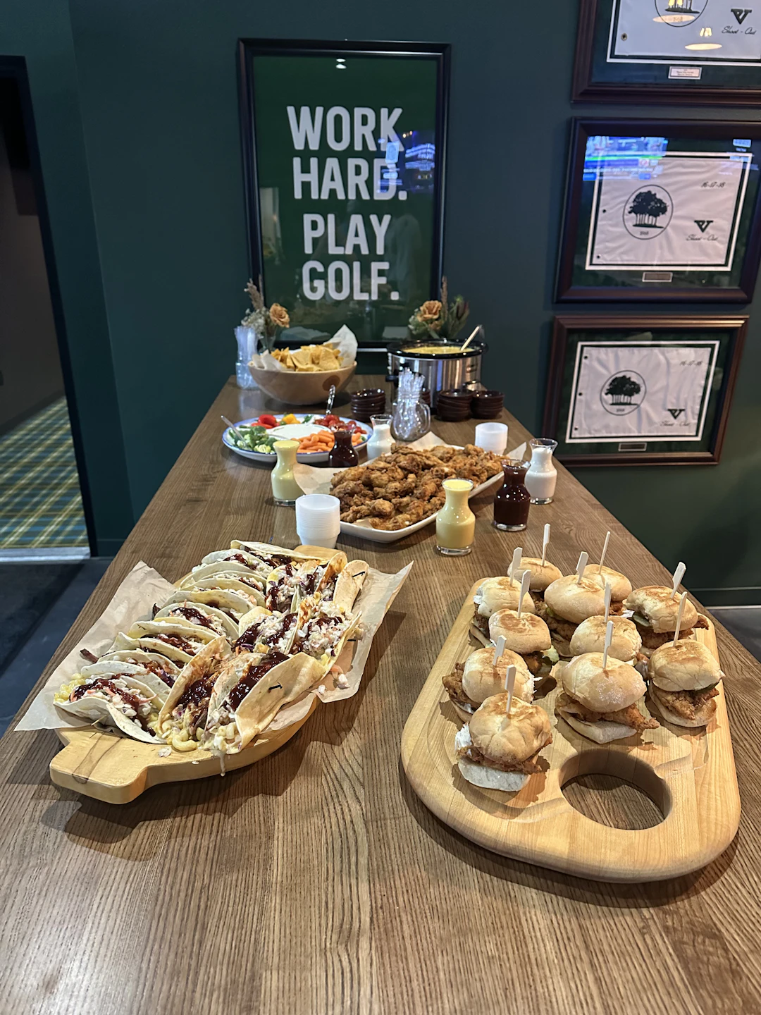 This image shows a wooden table set up with various food items. On the left side of the table, there is a platter of tacos with toppings and sauces. On the right side, there is a platter of sliders or mini burgers, each with a toothpick. In the center, there are several condiments and sauces, including what appears to be a creamy dip. Above the food, there is a framed sign with the words "WORK HARD. PLAY GOLF." mounted on the wall. Additionally, there are several framed certificates or awards displayed on the wall to the right of the sign.