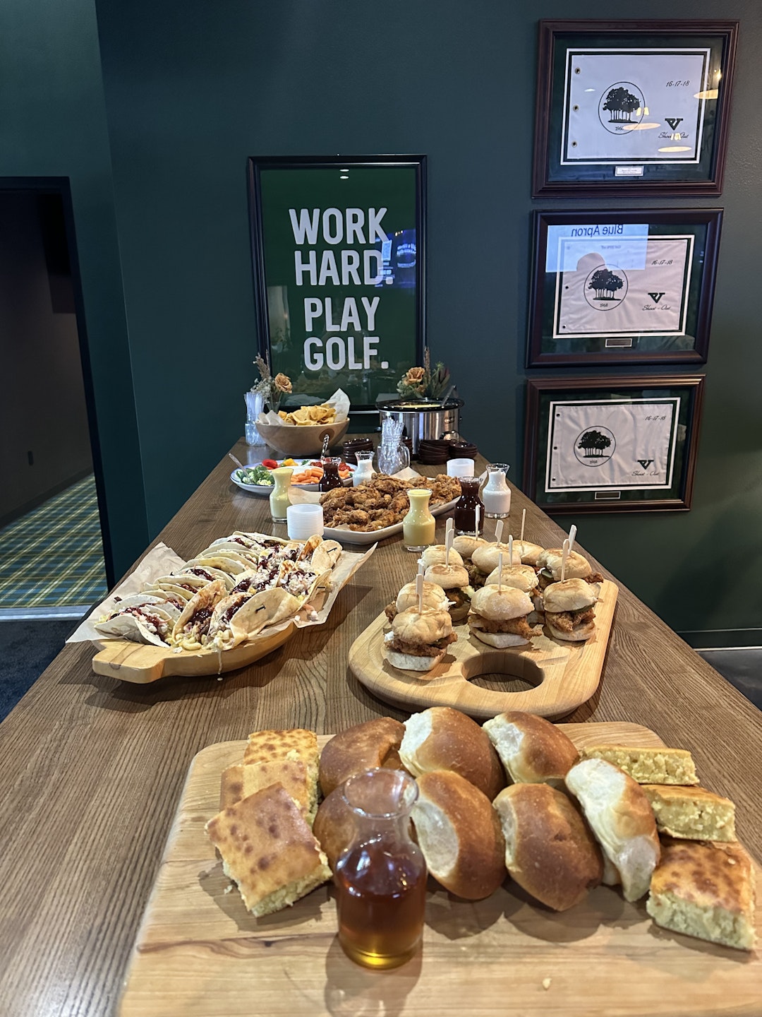 This image shows a wooden table set up with various food items. On the left side of the table, there is a platter of tacos with toppings and sauces. On the right side, there is a platter of sliders or mini burgers, each with a toothpick. In the center, there are several condiments and sauces, including what appears to be a creamy dip. Above the food, there is a framed sign with the words "WORK HARD. PLAY GOLF." mounted on the wall. Additionally, there are several framed certificates or awards displayed on the wall to the right of the sign.