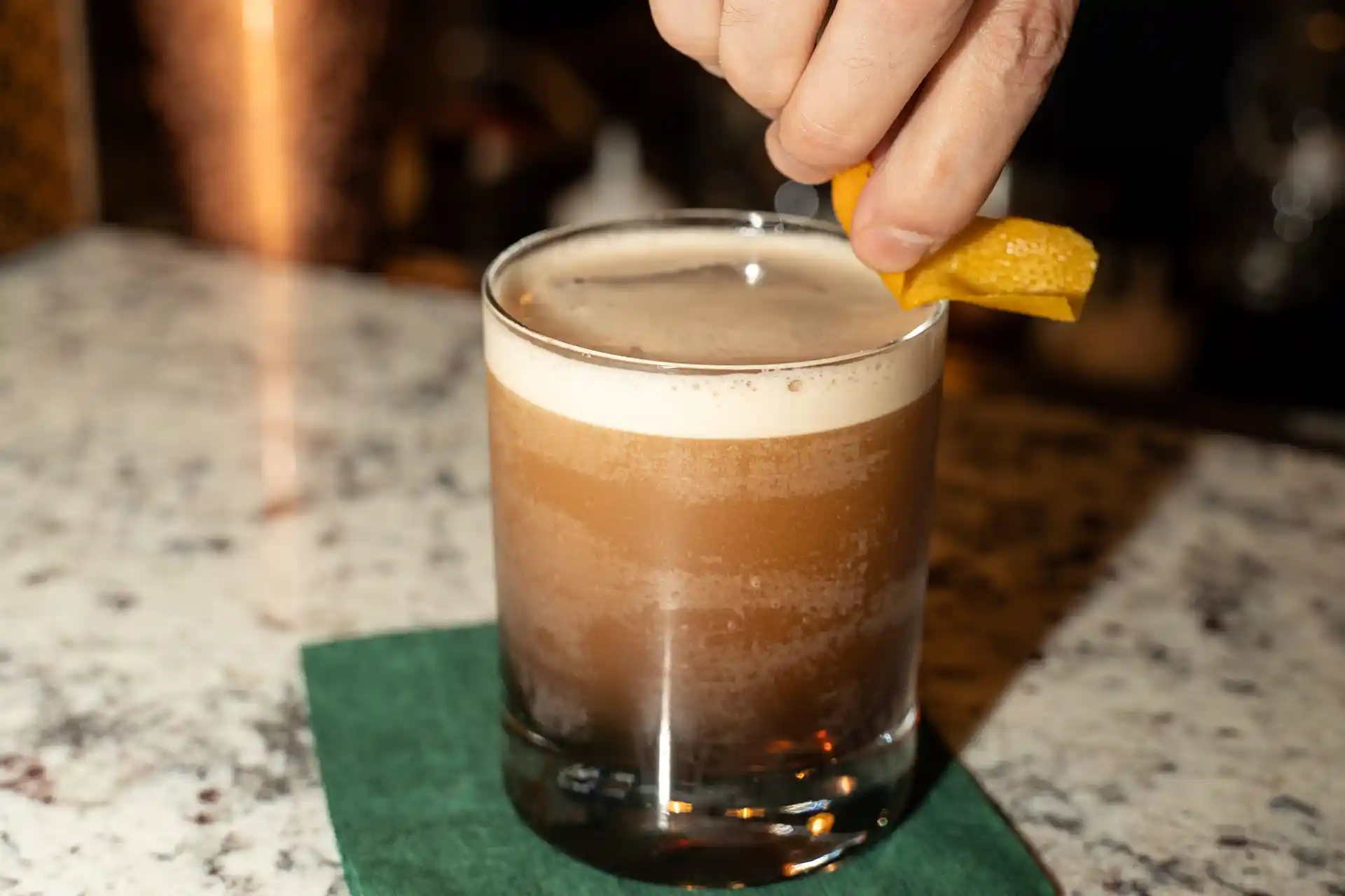A hand holds a twist of orange peel over a frothy brown cocktail in a glass, which sits on a green napkin on a speckled countertop.