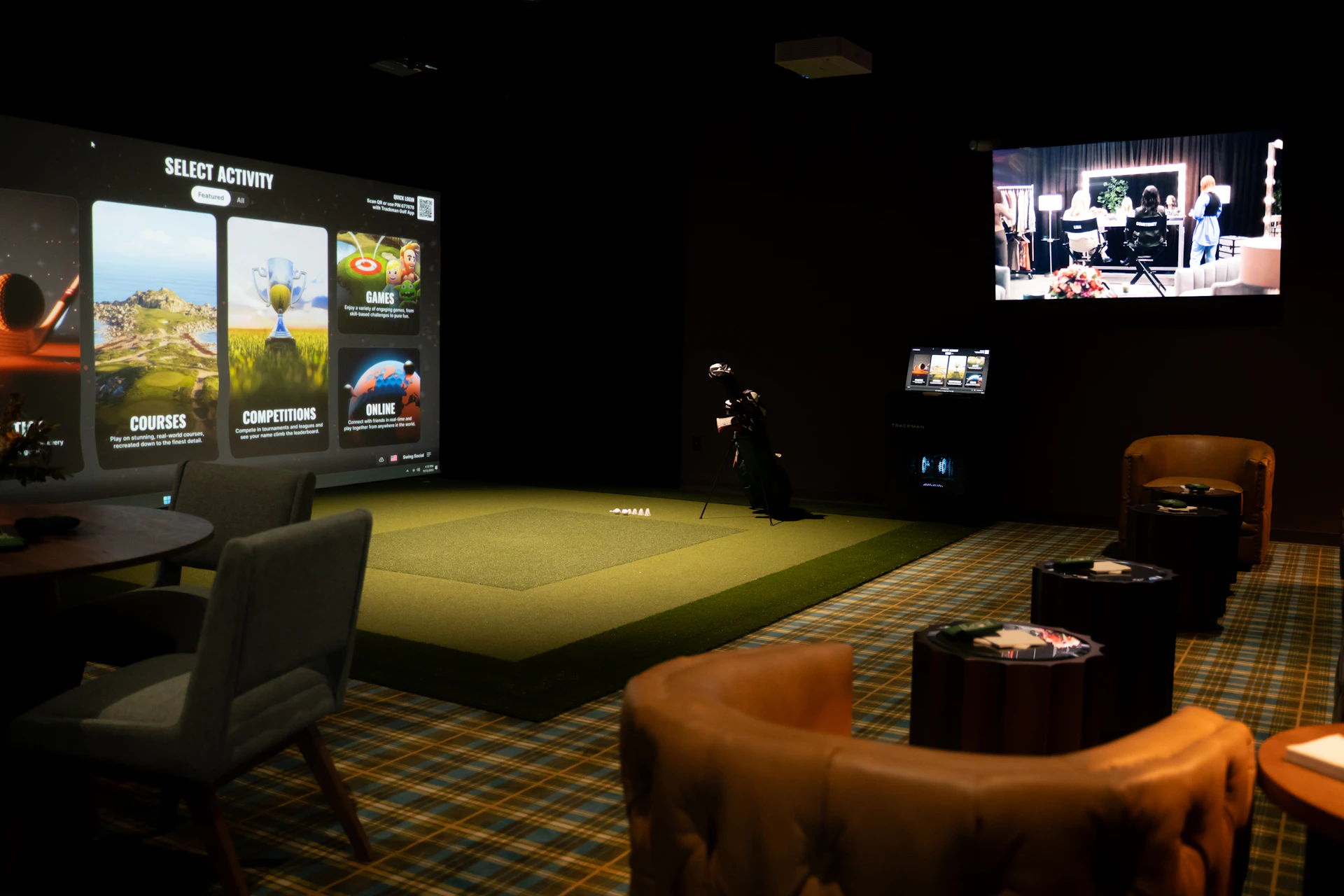 This image shows an indoor golf simulation room with a large projection screen displaying various golf-related activities such as courses, competitions, and games. The room has a green artificial turf area where a person is standing with a golf club, likely practicing or playing a simulated game. There are several chairs and tables arranged around the room, some with items on them, suggesting a comfortable and relaxed atmosphere. The walls are dark, enhancing the visibility of the projected images. The overall setup indicates a high-tech, recreational space designed for golf enthusiasts.