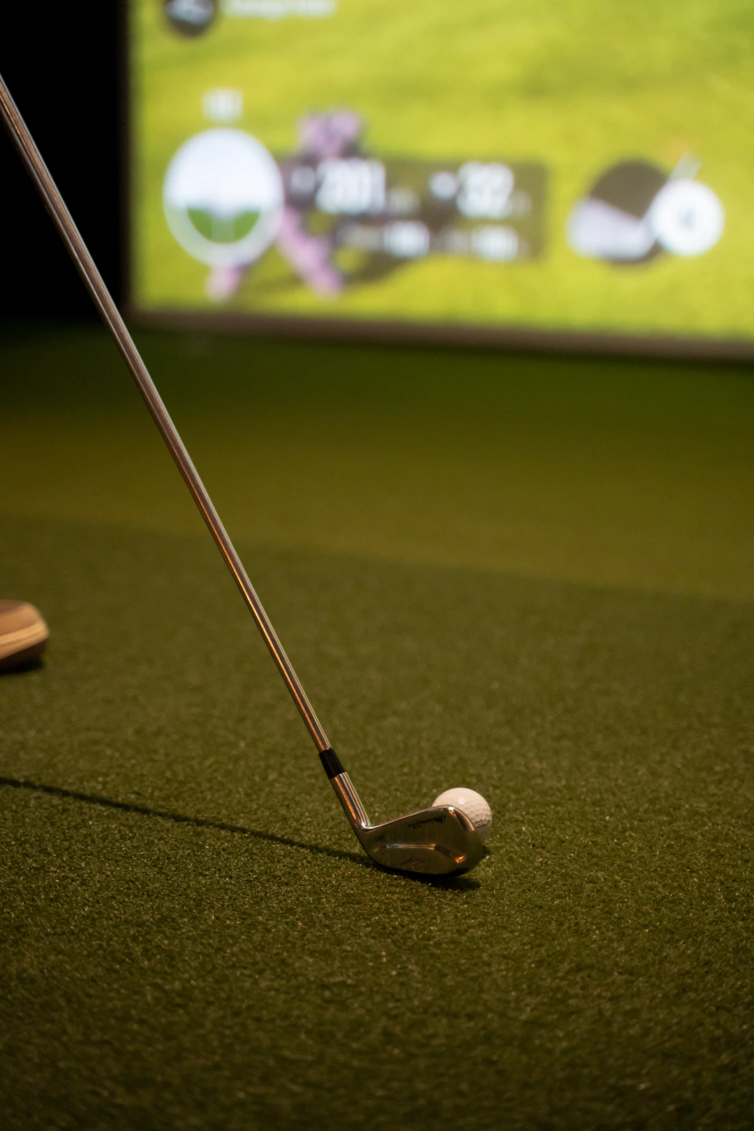 This image shows a close-up view of a golf club with a ball positioned on a green mat, likely in a golf driving range. The club is angled towards the ball, suggesting preparation for a swing. The background features a large screen displaying a golf-related graphic, including a silhouette of a golfer and some text, though the text is not clearly legible. The setting appears to be indoors, given the artificial green mat and the presence of the screen.