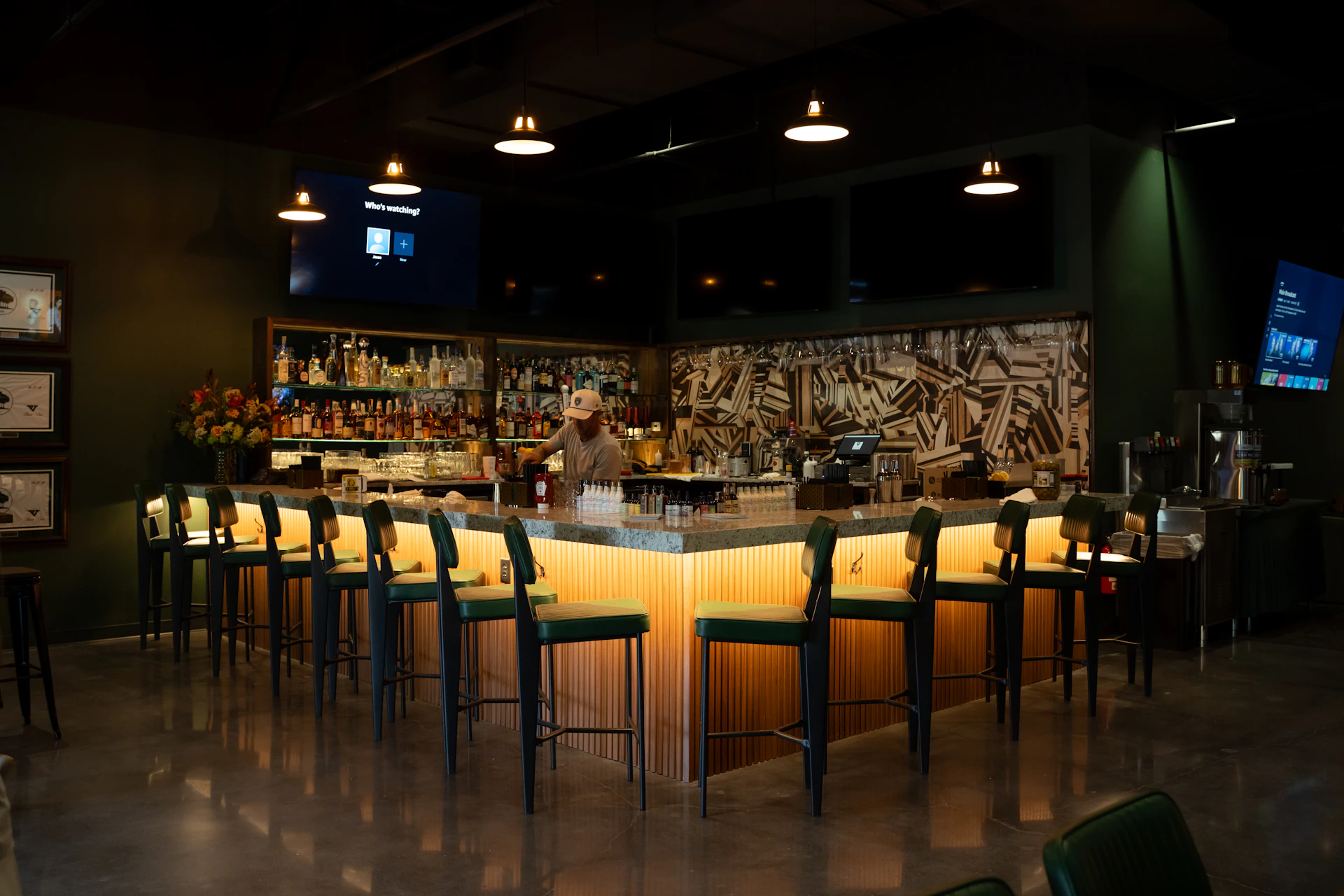 This image shows a person interacting with a digital display, likely a touchscreen, that lists various golf courses. The screen is divided into sections, each featuring a course name and a small image. The courses include names like "BANDON PRESERVE," "CABOT CITRUS FARMS," and "BETHPAGE BLACK." The person's hand is pointing at one of the courses, indicating an interaction or selection. The background is dimly lit, with a warm glow from a nearby lamp, creating a cozy atmosphere.