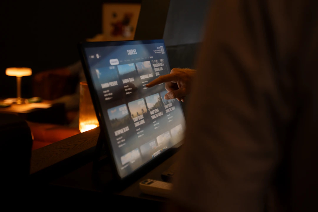 This image shows a person interacting with a digital display, likely a touchscreen, that lists various golf courses. The screen is divided into sections, each featuring a course name and a small image. The courses include names like "BANDON PRESERVE," "CABOT CITRUS FARMS," and "BETHPAGE BLACK." The person's hand is pointing at one of the courses, indicating an interaction or selection. The background is dimly lit, with a warm glow from a nearby lamp, creating a cozy atmosphere.