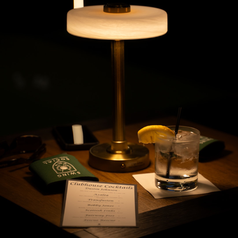 This image shows a dimly lit scene with a wooden table at the center. On the table, there is a lamp with a circular, frosted glass shade, casting a soft glow. Next to the lamp, there is a glass of cocktail with a lemon slice on top and a straw inside. A menu card titled "Clubhouse Cocktails" is placed on the table, listing various cocktail names such as "Azalea," "Transfusion," and "Fairway Fizz." Additionally, there is a green cloth with the logo and text "SNIMS" on it, placed near the menu card.