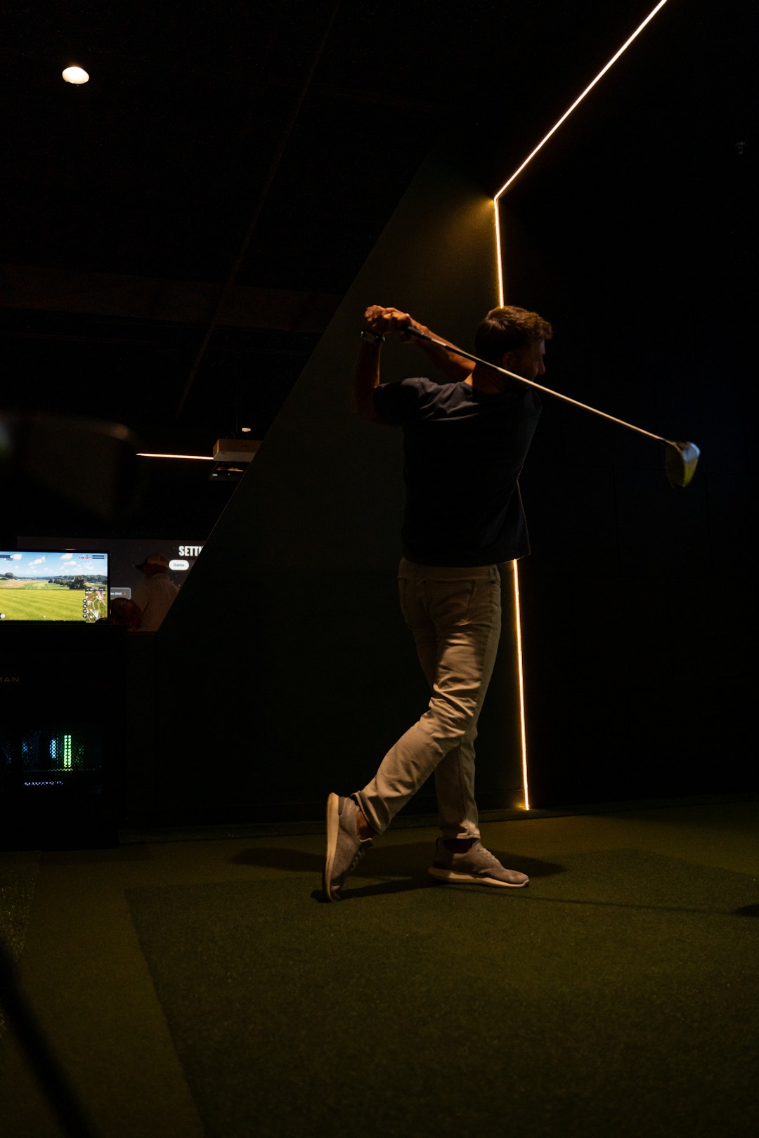 This image shows a person engaged in a golf swing within a dimly lit indoor golf facility. The individual is dressed in a dark-colored top and light-colored pants, with a golf club held firmly in both hands. The background features a large screen displaying a golf course, suggesting a virtual golfing environment. The screen shows a green field with a golfer, indicating a simulated golfing experience. The facility is illuminated by vertical lines of light on the walls, creating a focused ambiance on the golfer. The word "SETT" appears multiple times on the screen, possibly indicating the name of the golf simulation software or facility.