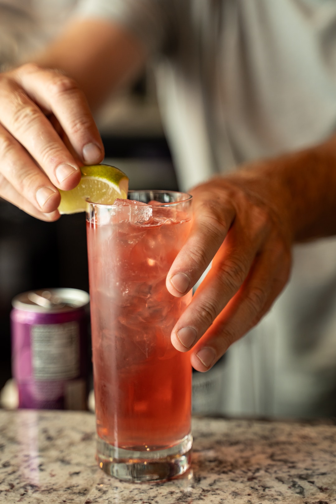 This image shows a person preparing a drink by adding a slice of lime to a glass filled with a red beverage and ice cubes. The glass is placed on a marble surface. In the background, there is a can of a beverage, which appears to be a soft drink, with a purple label. The person's hands are the main focus, with one hand holding the lime and the other holding the glass.