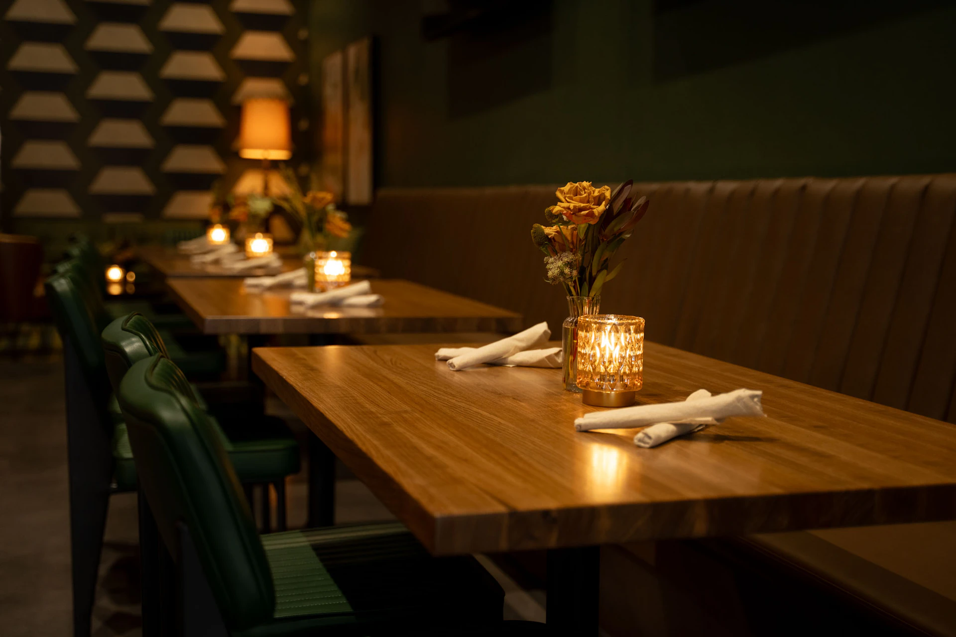 This image shows an elegant dining area with wooden tables and green cushioned chairs. Each table is set with white napkins, small lit candles, and a vase with flowers. The tables are arranged in a row along a cushioned bench seating area. The ambiance is warm and inviting, with soft lighting from the candles and a lamp in the background. The walls feature geometric patterns and framed artwork, contributing to the sophisticated decor of the restaurant.