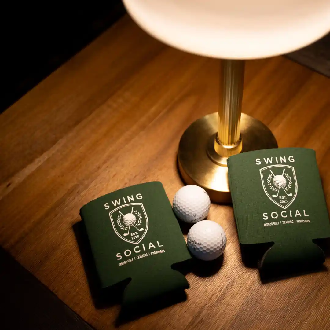 Two green drink koozies labeled Swing Social with golf logos sit on a wooden table next to two white golf balls and a brass lamp with a white shade.