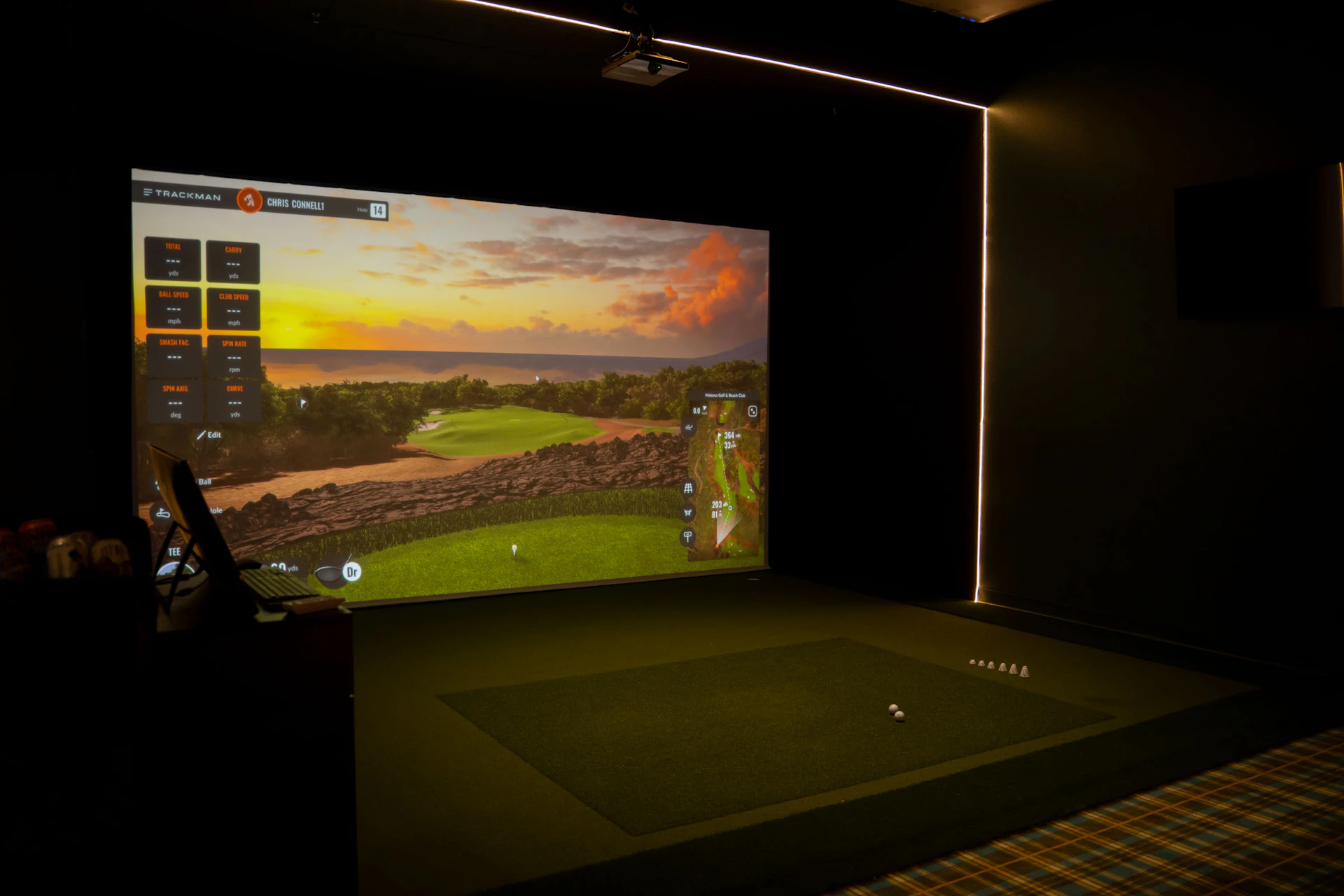 This image shows a golf simulation room with a large screen displaying a virtual golf course at sunset. The screen shows various statistics and metrics related to a golf shot, such as ball speed, club speed, spin rate, and carry distance. The room has a green artificial turf area where golf balls are placed, and there is a computer monitor on the left side likely used for controlling the simulation. The walls are dark, and there are illuminated lines on the ceiling, enhancing the ambiance of the simulation environment.