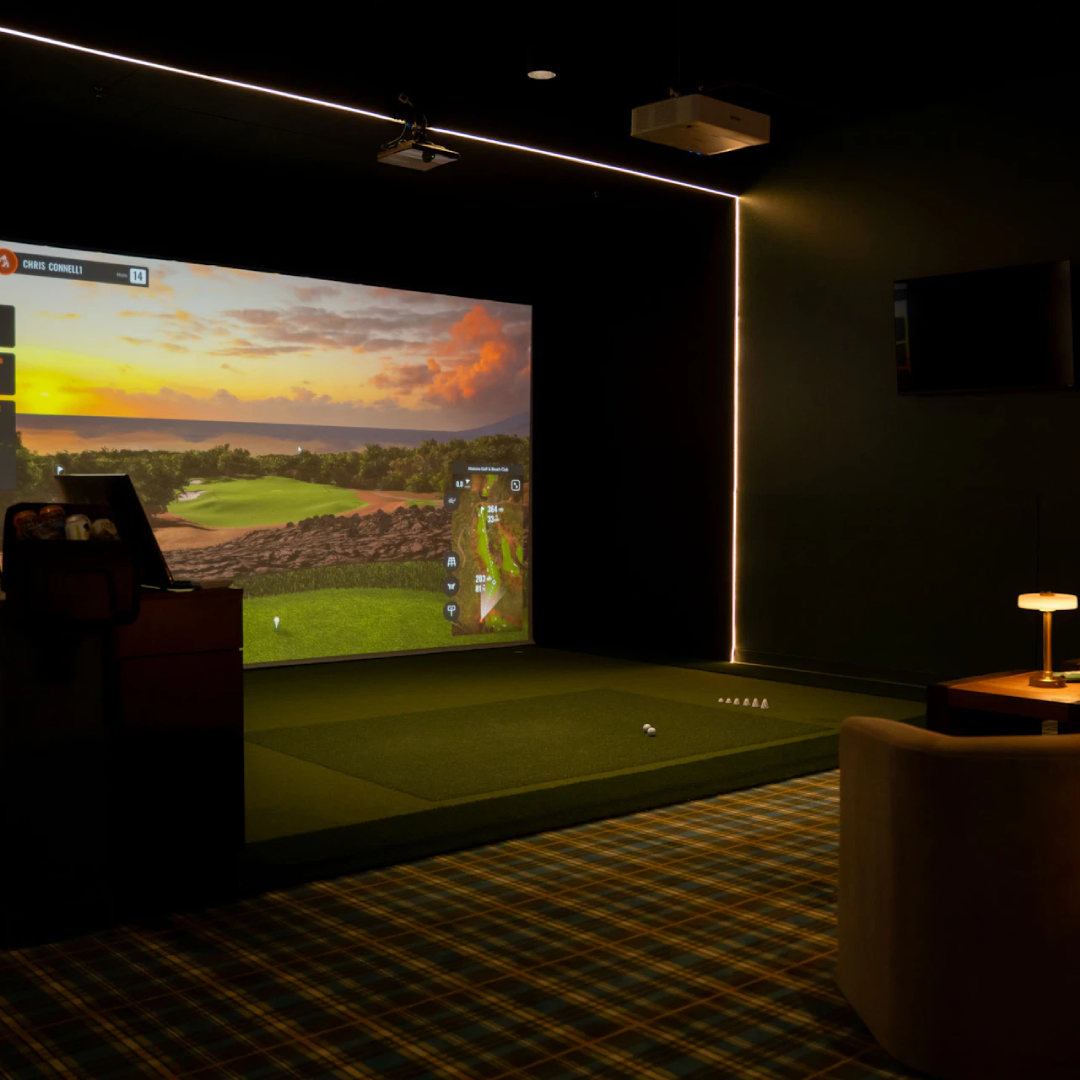 This image shows a person engaged in a golf activity in a dimly lit indoor setting. The person is bent over, holding a golf club, and appears to be preparing to hit a golf ball. In the background, there is a large screen displaying a virtual golf course with various statistics and labels such as "DEEP ROUGH," "249 yds," and "TRACKMAN." The screen also shows yardages and other golfing metrics. The person is wearing a dark shirt and light-colored pants.