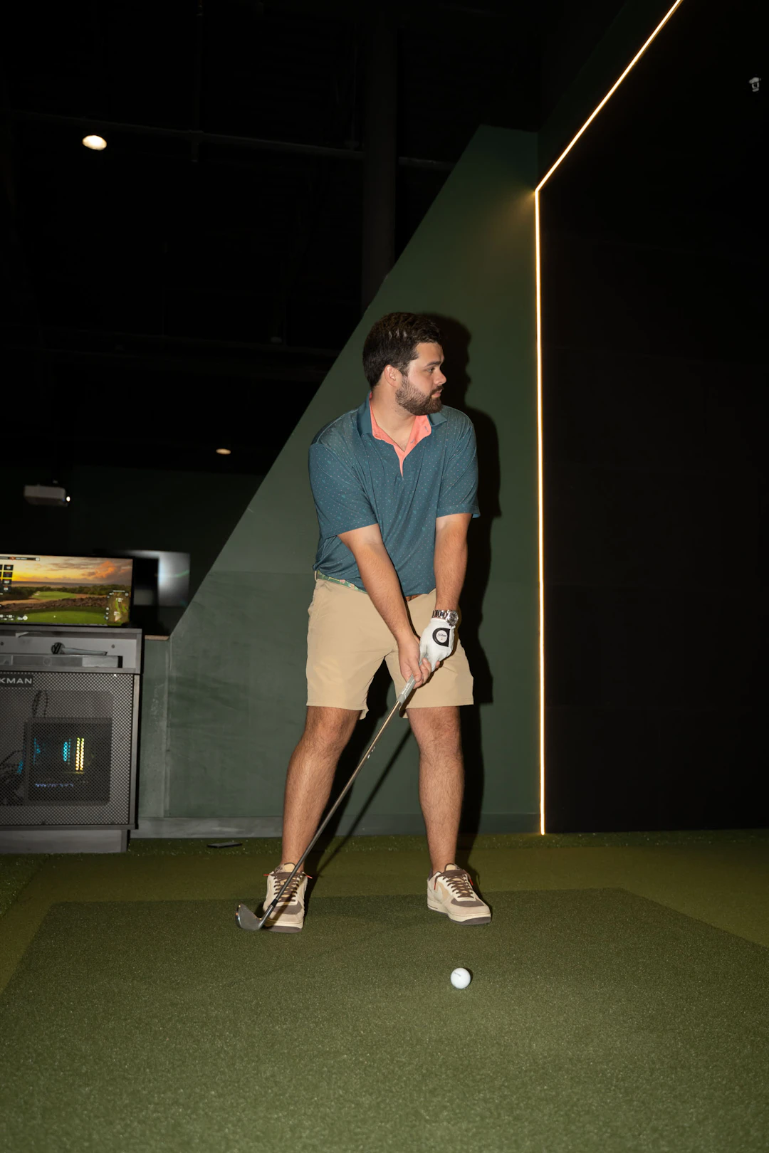 A man stands indoors on artificial turf, holding a golf club and preparing to swing. He is wearing a teal polo shirt, khaki shorts, and sneakers, and is looking to his left. A golf simulator is visible in the background.