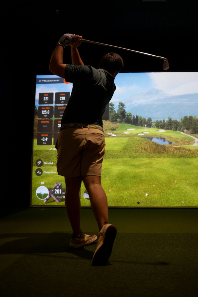 This image shows a person engaged in a golf swing activity in a simulated environment. The individual is holding a golf club and appears to be hitting a golf ball. In the background, there is a large screen displaying various statistics and information related to the golf swing, such as ball speed, carry distance, and spin axis. The screen also shows a virtual golf course with lush green fields, trees, and a distant mountain range. The person is dressed in casual attire, including shorts and a t-shirt, suitable for indoor sports activities.