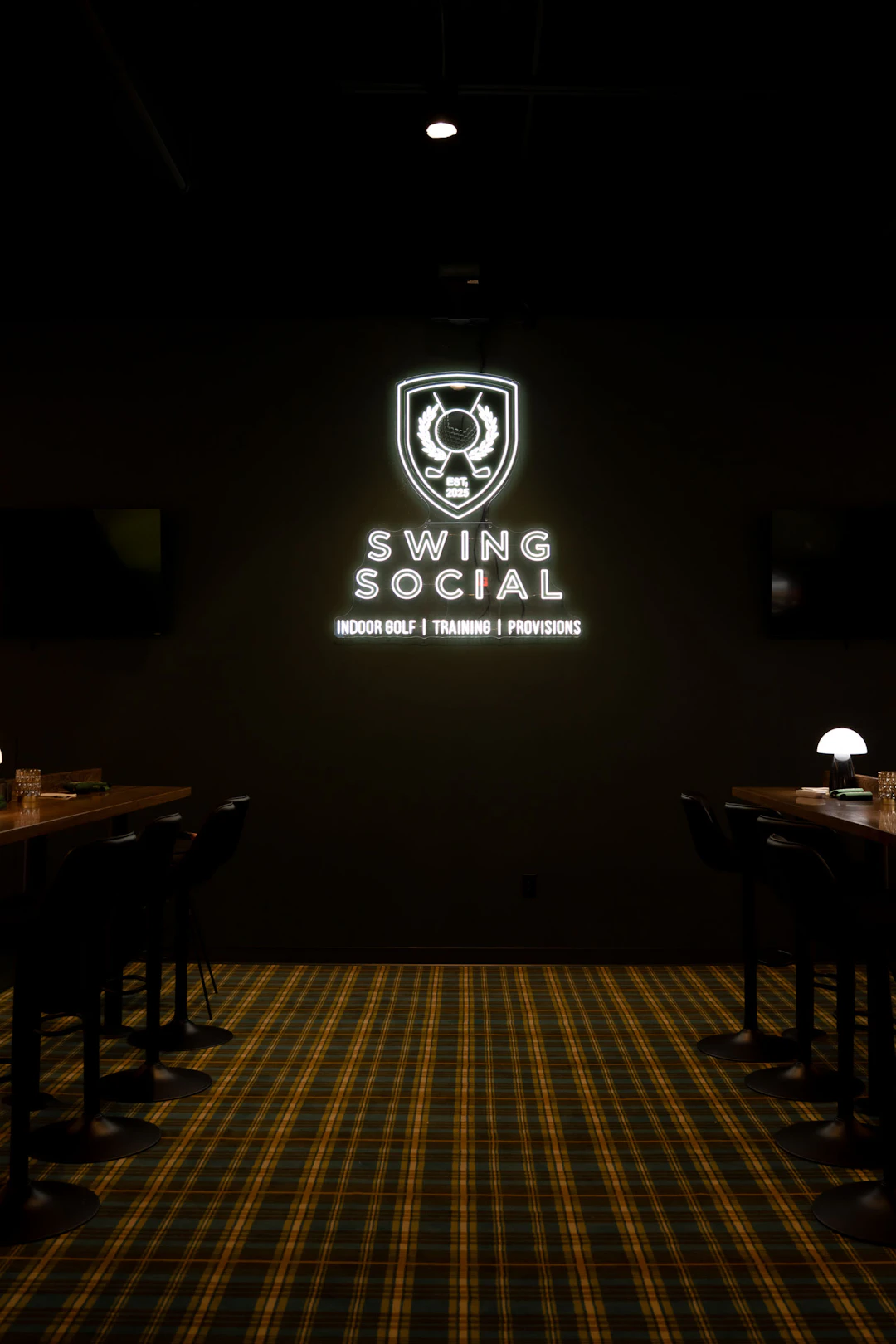 This image shows an indoor space with a dark ambiance, illuminated by a prominent neon sign on the wall that reads "SWING SOCIAL" with a logo featuring a golf ball and clubs. Below the neon sign, there is additional text that reads "INDOOR GOLF | TRAINING | PROVISIONS". The room has a patterned carpet with a mix of yellow and blue colors. There are tables with chairs arranged along the sides of the room, and a small lamp is visible on one of the tables. The overall setting suggests a venue for indoor golfing activities and social gatherings.