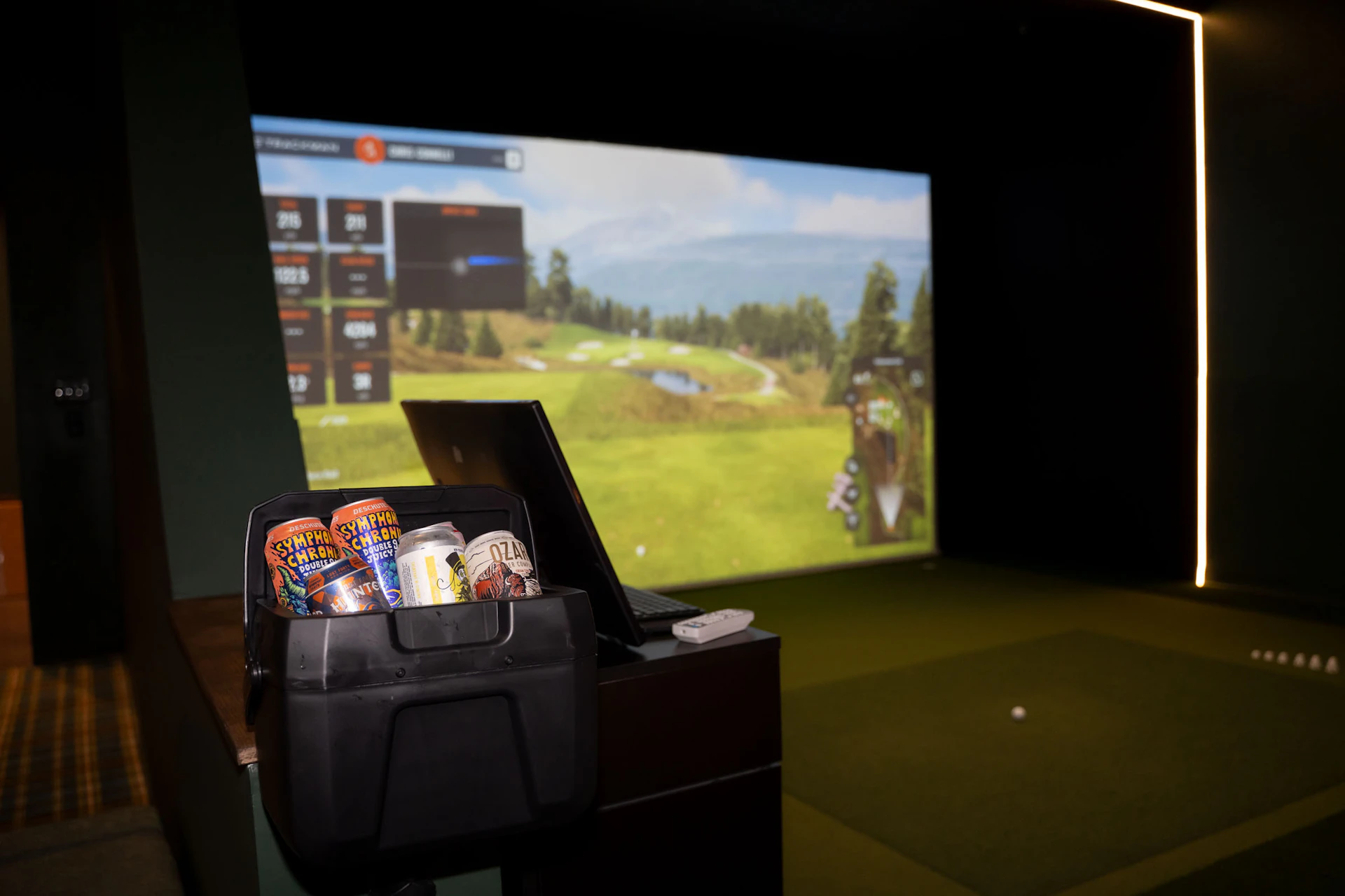 This image shows a golf simulation setup with a large screen displaying a virtual golf course. In front of the screen, there is a black cooler containing several cans of Deschutes Brewery beer, including Symphony Chromatic Double IPA and Juic DIPA. Next to the cooler, there is a laptop open on a desk, and a remote control is placed nearby. The area is dimly lit with a yellow light strip on the right side of the screen.