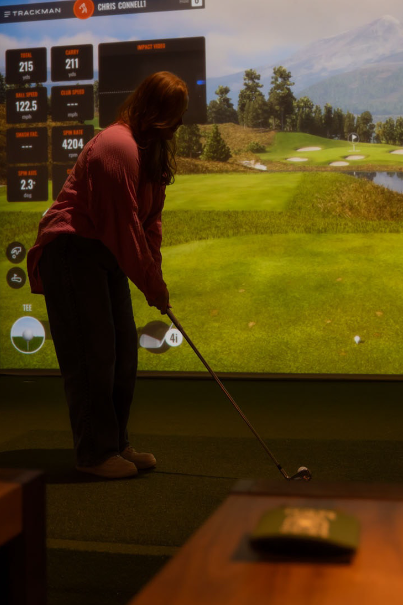This image shows a person engaged in a golf simulation activity in a dimly lit room. The person is standing in front of a large screen displaying a virtual golf course, holding a golf club, and appears to be preparing to hit a ball. The screen shows various statistics and details about the player's performance, including scores and metrics such as club speed and ball speed. There are two tables with lamps on either side of the person, providing additional lighting. The overall setup suggests a high-tech, immersive golfing experience.