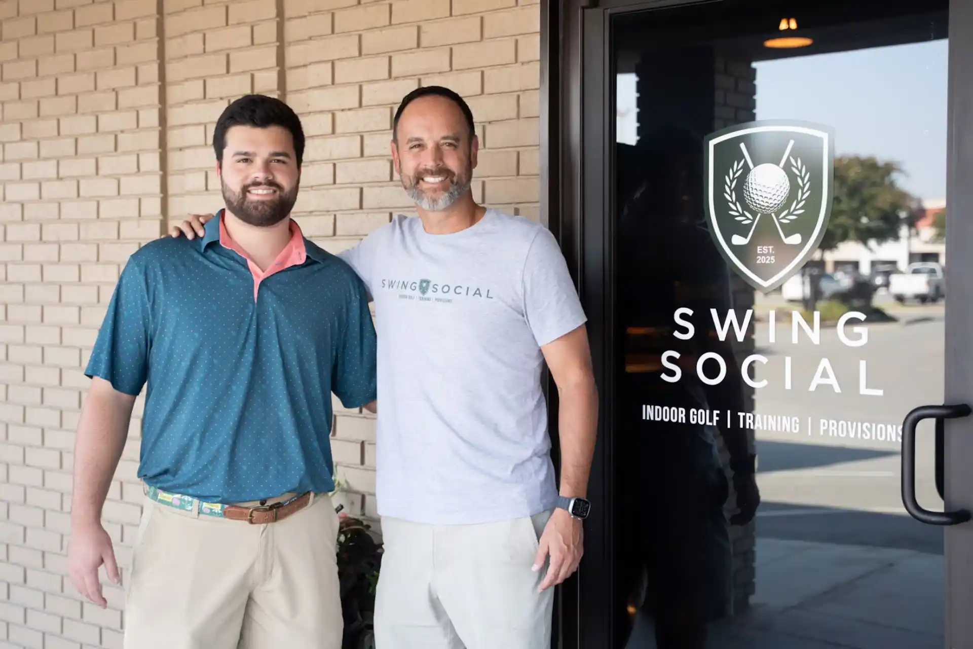 This image shows two individuals standing next to each other in front of a building. The building has a glass door with a logo and text on it. The logo features a golf ball and clubs, and the text reads "SWING SOCIAL" along with additional information about indoor golf, training, and provisions. The person on the left is wearing a blue polo shirt with a pink collar and khaki pants, while the person on the right is wearing a gray t-shirt and white pants. Both individuals appear to be smiling and posing for a photograph.
