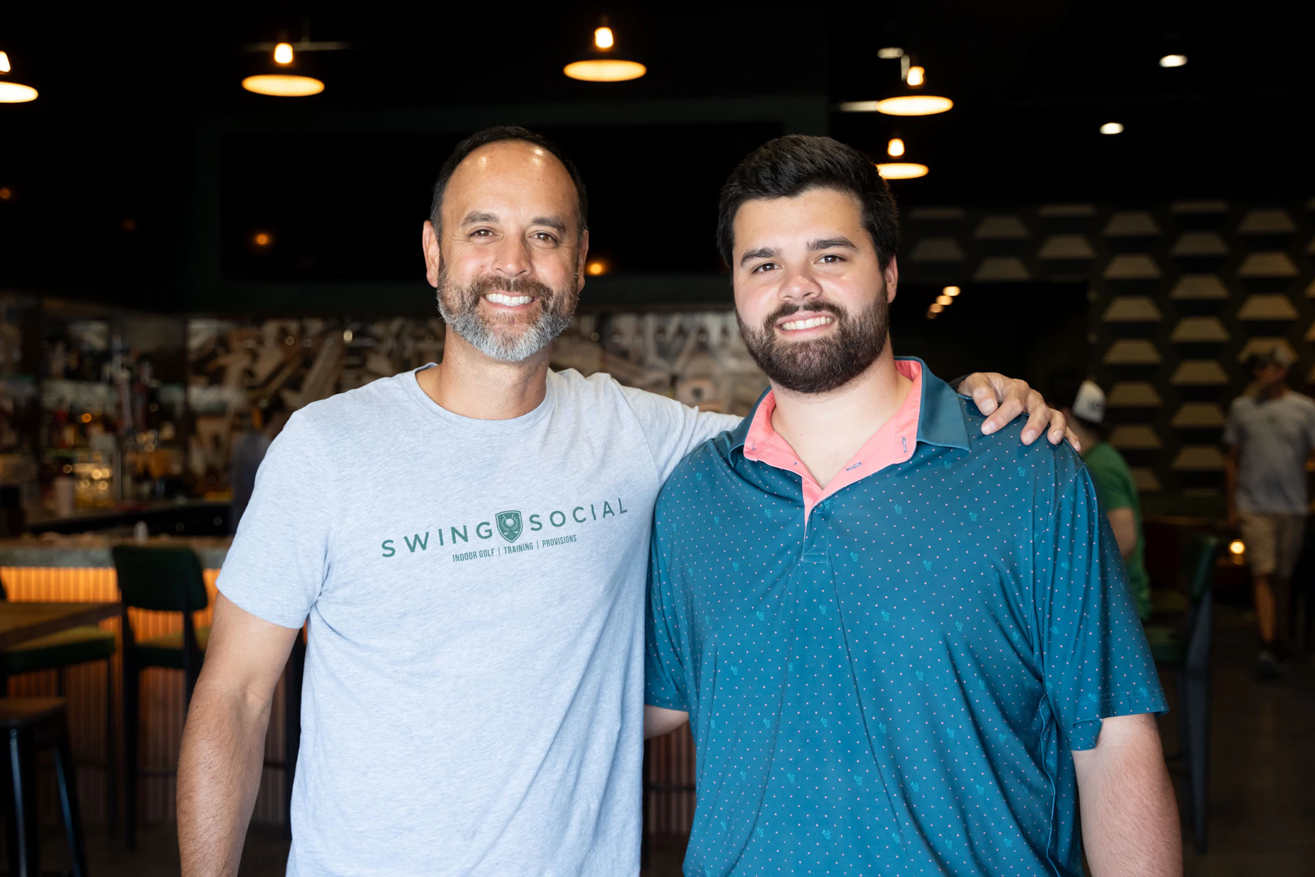 This image shows two individuals standing close to each other, smiling and posing for a photograph. The person on the left is wearing a light gray t-shirt with the text "SWING SOCIAL" and a logo on it. The person on the right is wearing a dark blue polo shirt with white dots. Behind them, there is a bar counter with various items on display and several people in the background, suggesting a social or recreational setting. The lighting is dim with several hanging lights providing illumination.