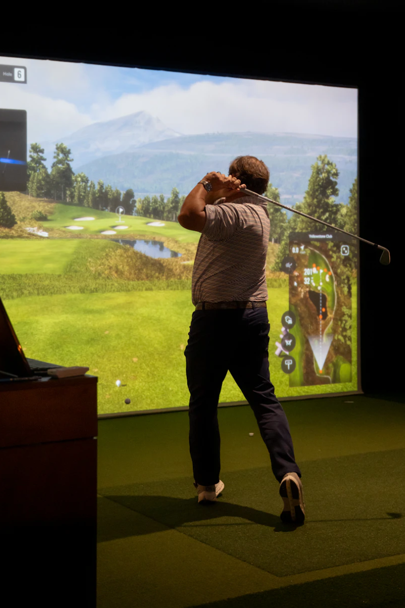 This image shows a person engaged in a golf driving range activity indoors. The individual is holding a golf club and appears to be swinging it, with their body positioned in a typical golf stance. In the background, there is a large screen displaying a virtual golf course with lush green fields, trees, and a distant mountain range under a clear sky. The screen also shows various texts and numbers, likely indicating details about the golf course or the player's performance, such as hole numbers and scores. The setting is well-lit with artificial lighting, and the floor is covered with artificial turf.