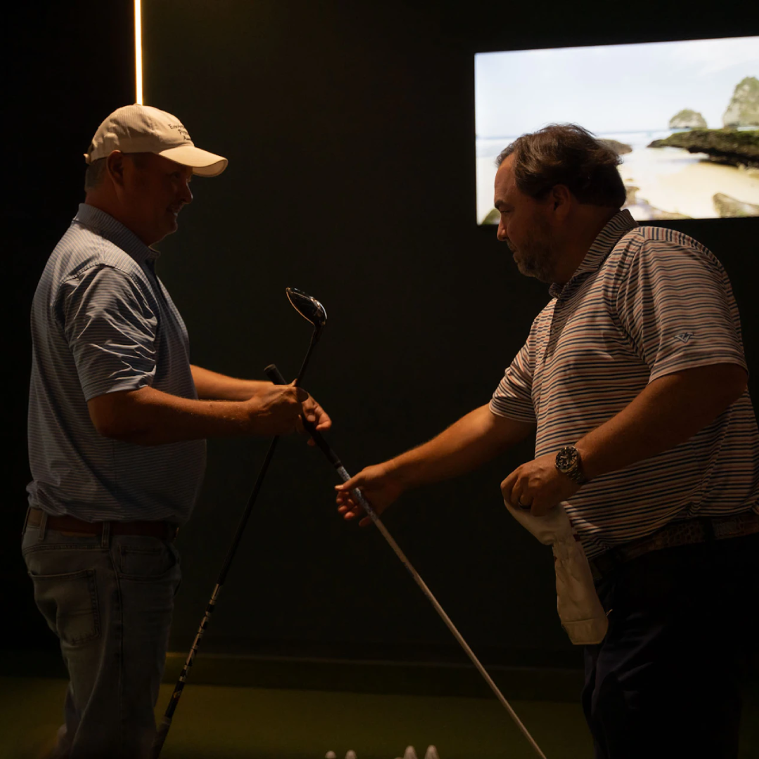This image shows two men standing in a dimly lit indoor area, possibly a golf practice range. The man on the left is holding a golf club and appears to be demonstrating or explaining something to the man on the right, who is also holding a golf club. The man on the right is wearing a watch and holding a white cloth. Behind them, there is a large screen displaying a scenic outdoor view, likely used for simulating different golfing environments. The overall setting suggests a focused and instructional atmosphere.