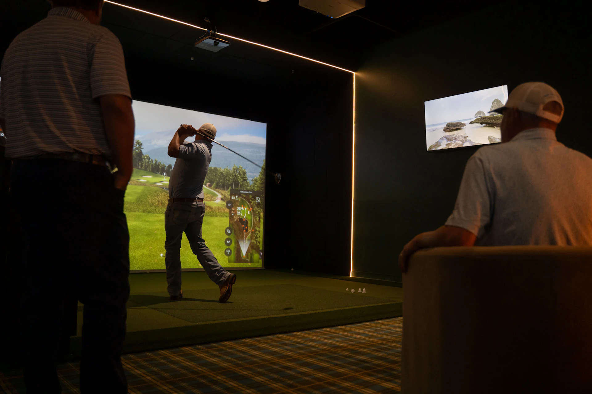 This image shows a group of people in a golf driving range. One person is actively swinging a golf club, while others are observing. The background features a large screen displaying a golf course scene. The room is dimly lit with a few lights accentuating the area. The floor has a green mat for the golfer to stand on, and there are golf balls scattered around. The observer on the right is seated in a chair, watching the golfer.