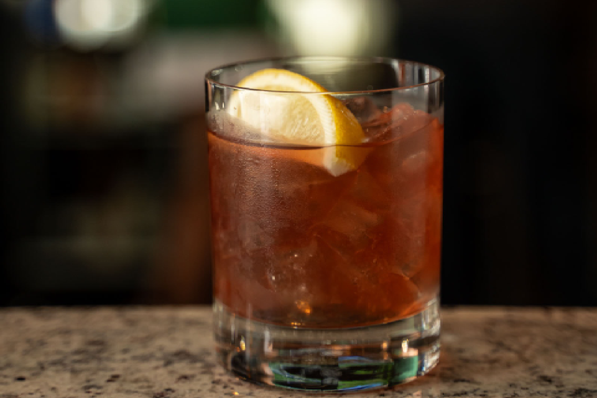 This image shows a glass of a dark-colored beverage, likely an alcoholic cocktail, placed on a marble surface. The drink appears to be a mix of various ingredients, possibly including whiskey or rum, with a slice of lemon on top. The background is blurred, showing a person standing near a green object, possibly a container or piece of equipment. The setting suggests a bar or a similar environment where drinks are served.