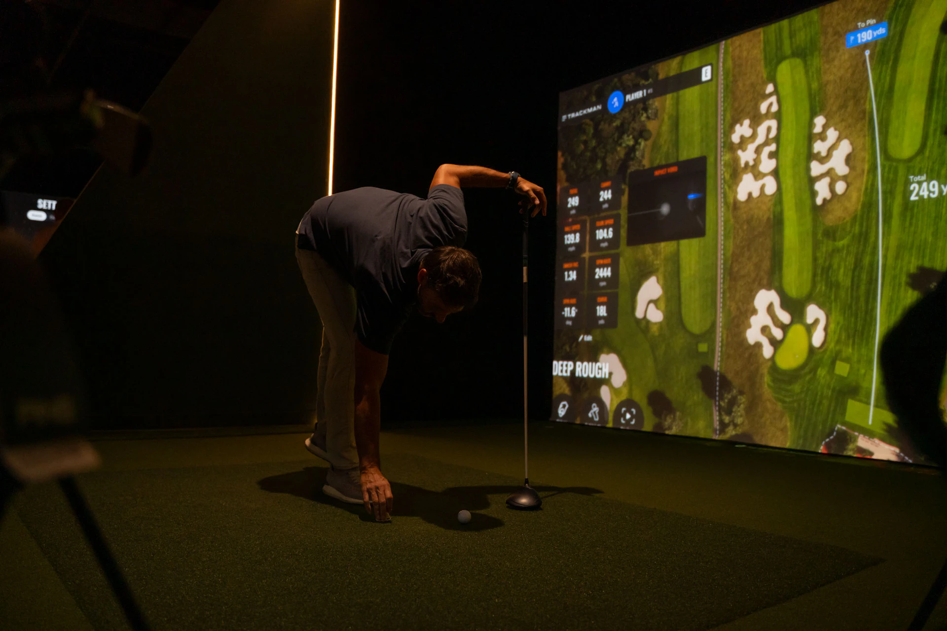 This image shows a person engaged in a golf activity in a dimly lit indoor setting. The person is bent over, holding a golf club, and appears to be preparing to hit a golf ball. In the background, there is a large screen displaying a virtual golf course with various statistics and labels such as "DEEP ROUGH," "249 yds," and "TRACKMAN." The screen also shows yardages and other golfing metrics. The person is wearing a dark shirt and light-colored pants.