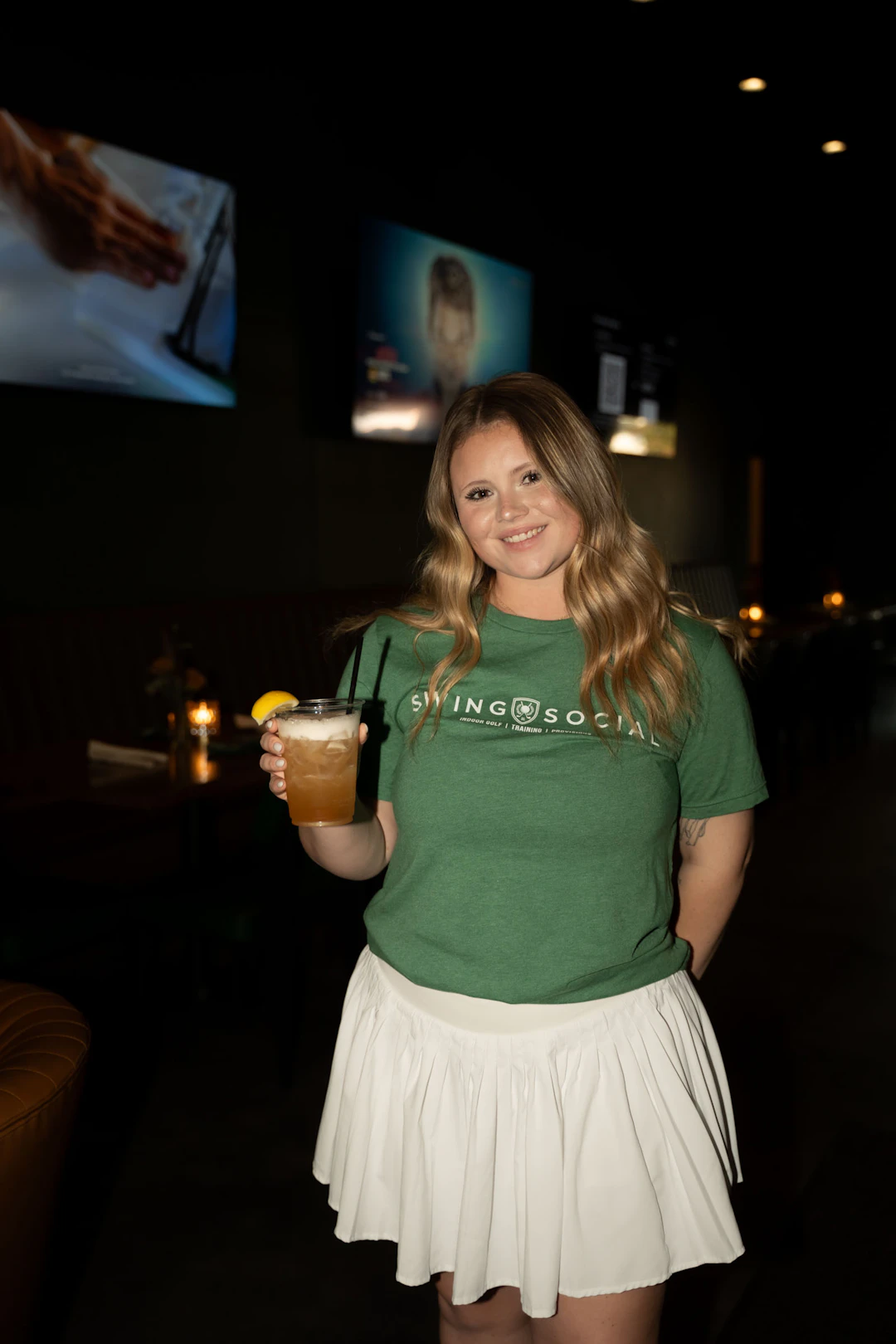A smiling woman in a green Swing Social t-shirt and white skirt holds a cocktail with a lemon garnish in a dimly lit bar, with TV screens and candles visible in the background.