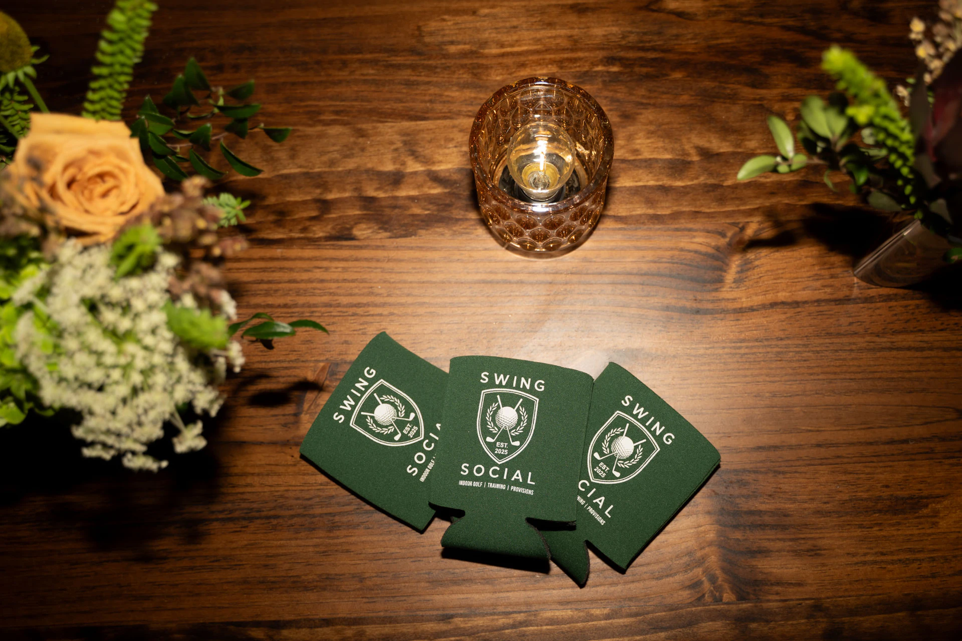 This image shows a wooden table with various items arranged on it. There are three green drink koozies with the text "SWING SOCIAL" and a logo featuring a golf ball and club. To the left, there is a floral arrangement with roses and greenery. On the right, there is another floral arrangement. In the center, there is a small glass candle holder with a lit candle inside. The overall setting appears to be a decorative and possibly social gathering setup.