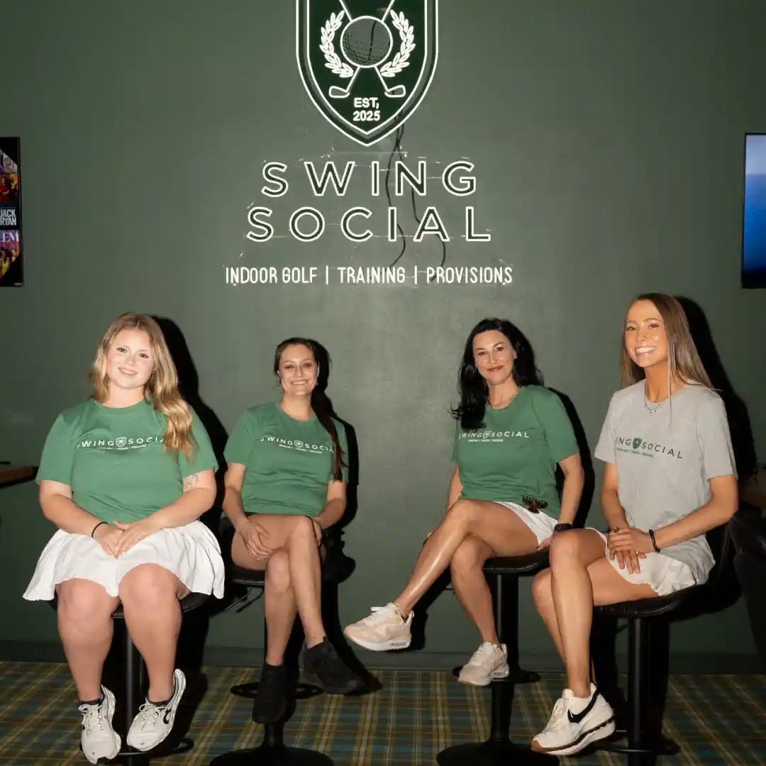 This image shows four individuals seated on stools in a room with a green wall. They are all wearing matching green t-shirts with the text "SWING SOCIAL" and white skirts. The wall behind them features a large logo with a golf emblem and the text "SWING SOCIAL" along with "INDOOR GOLF | TRAINING | PROVISIONS". There is also a smaller logo with the text "EST, 2025" above the main text. The floor has a plaid pattern, and the overall setting appears to be a promotional or team photo for a golf-related social club or service.