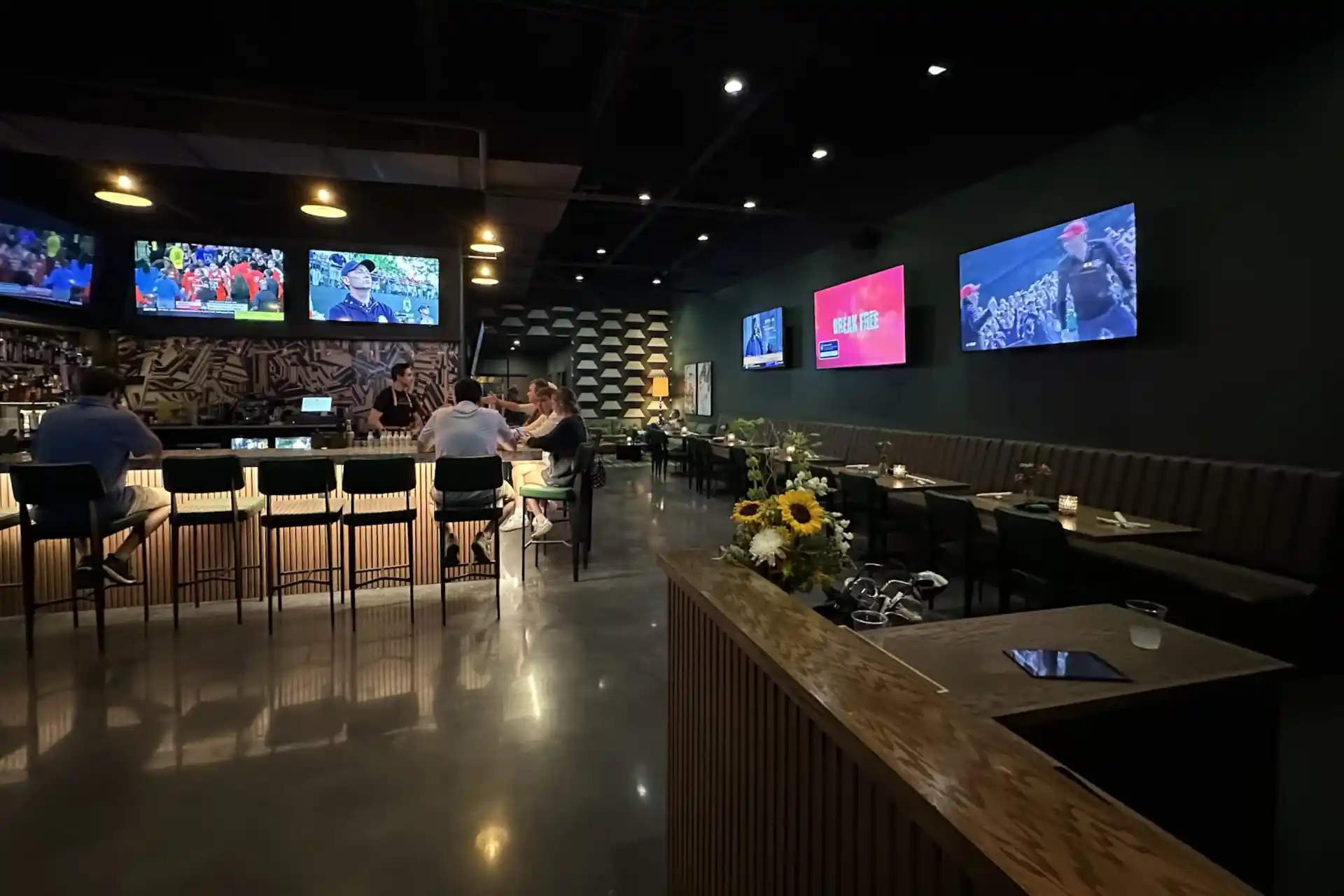 This image shows an indoor golf simulation room with a large projection screen displaying various golf-related activities such as courses, competitions, and games. The room has a green artificial turf area where a person is standing with a golf club, likely practicing or playing a simulated game. There are several chairs and tables arranged around the room, some with items on them, suggesting a comfortable and relaxed atmosphere. The walls are dark, enhancing the visibility of the projected images. The overall setup indicates a high-tech, recreational space designed for golf enthusiasts.